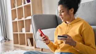 Young adult African American female consumer holding credit card and smartphone sitting on floor at home doing online banking transaction. E commerce virtual shopping, secure mobile banking concept.; Shutterstock ID 1979496134