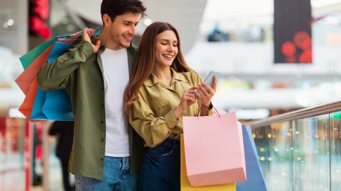 Buyers Couple Shopping Using Cellphone Holding Colorful Shopper Bags Standing In Mall. Happy Customers Using Application Purchasing Clothes Online Via Smartphone. Ecommerce And Shopaholism; Shutterstock ID 2116844222